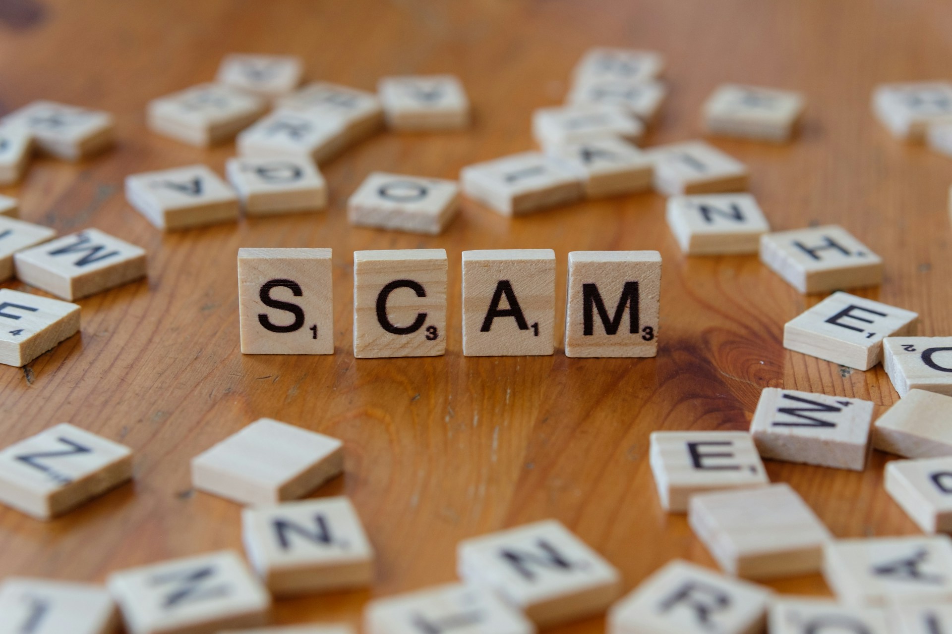 Wooden tiles spelling "SCAM" arranged among scattered alphabet letters on a wooden surface.