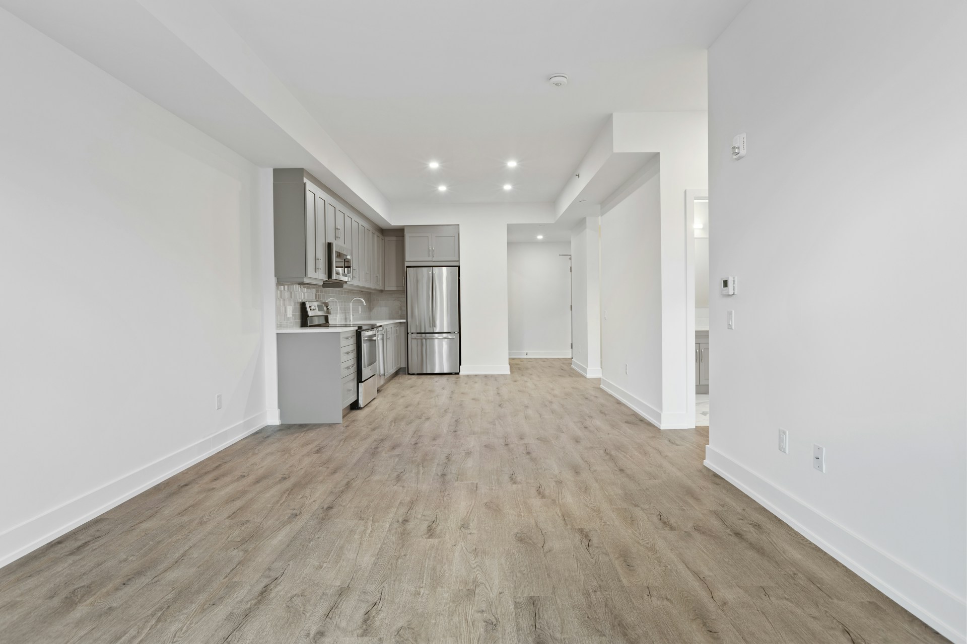 Bright, empty apartment interior with wood flooring, white walls, and kitchen visible in background hallway.