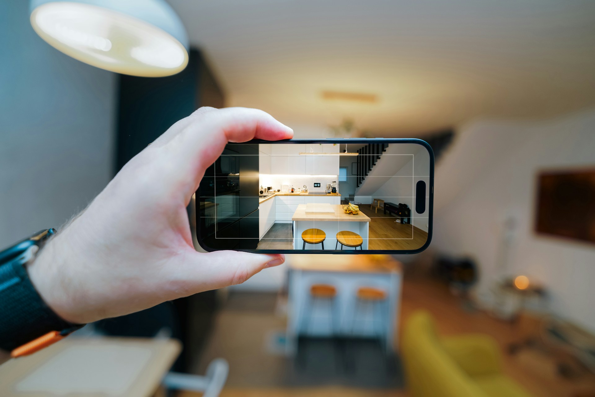 Hand holding smartphone photographing a bright, warmly-lit residential interior with furniture and doorway visible.