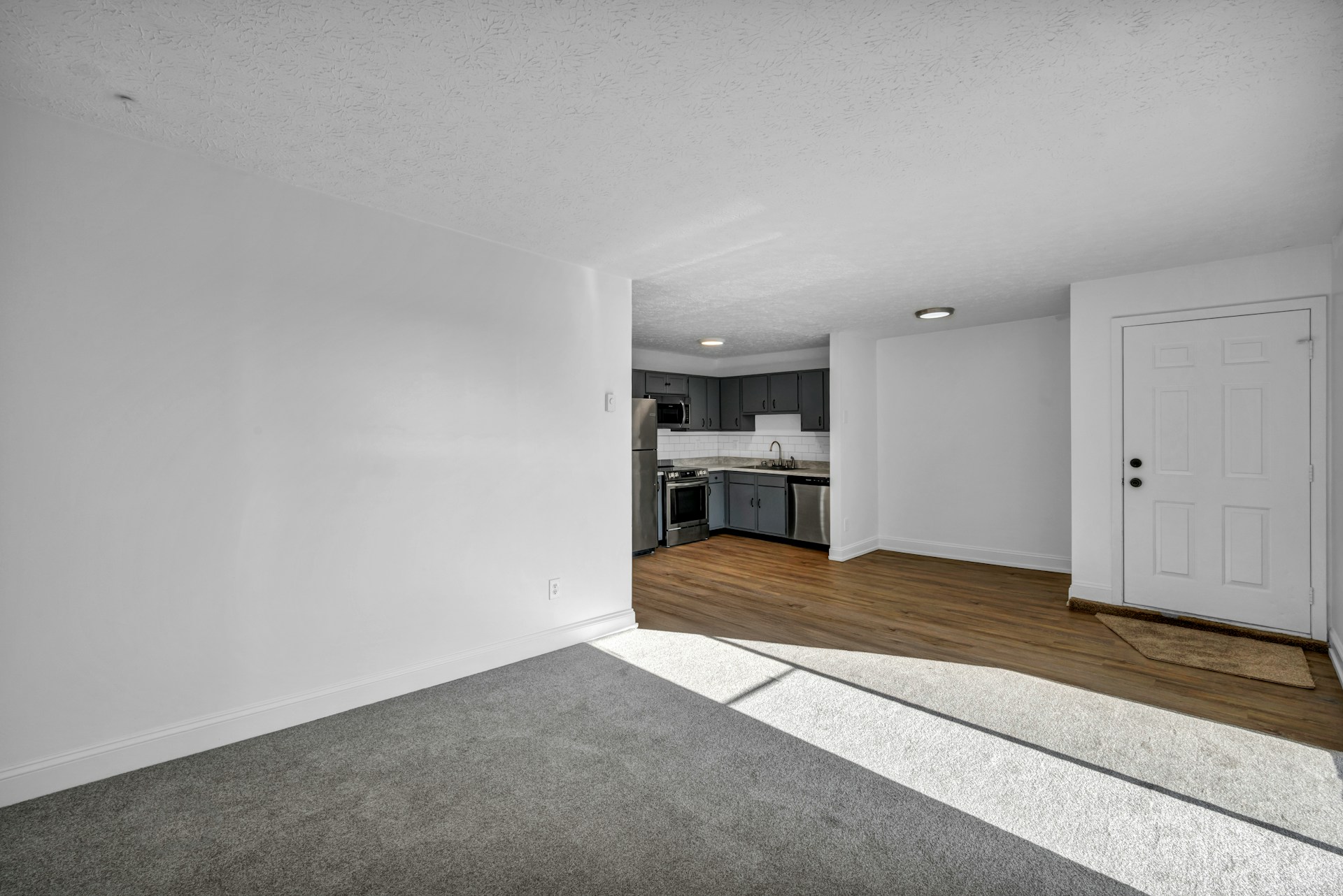 Vacant unit with grey carpet opening onto a kitchen with dark cabinets, between two tenancies.