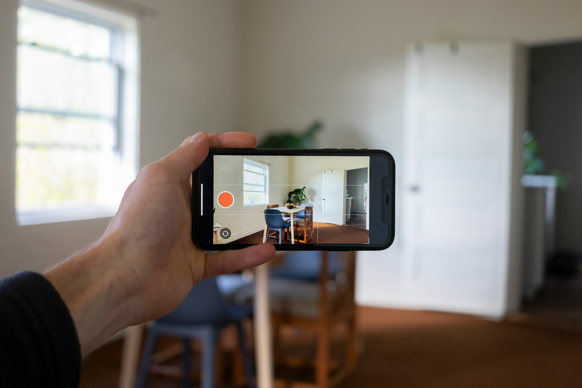 Person holding smartphone photographing a bright, empty residential room with white walls and wooden flooring during daytime.