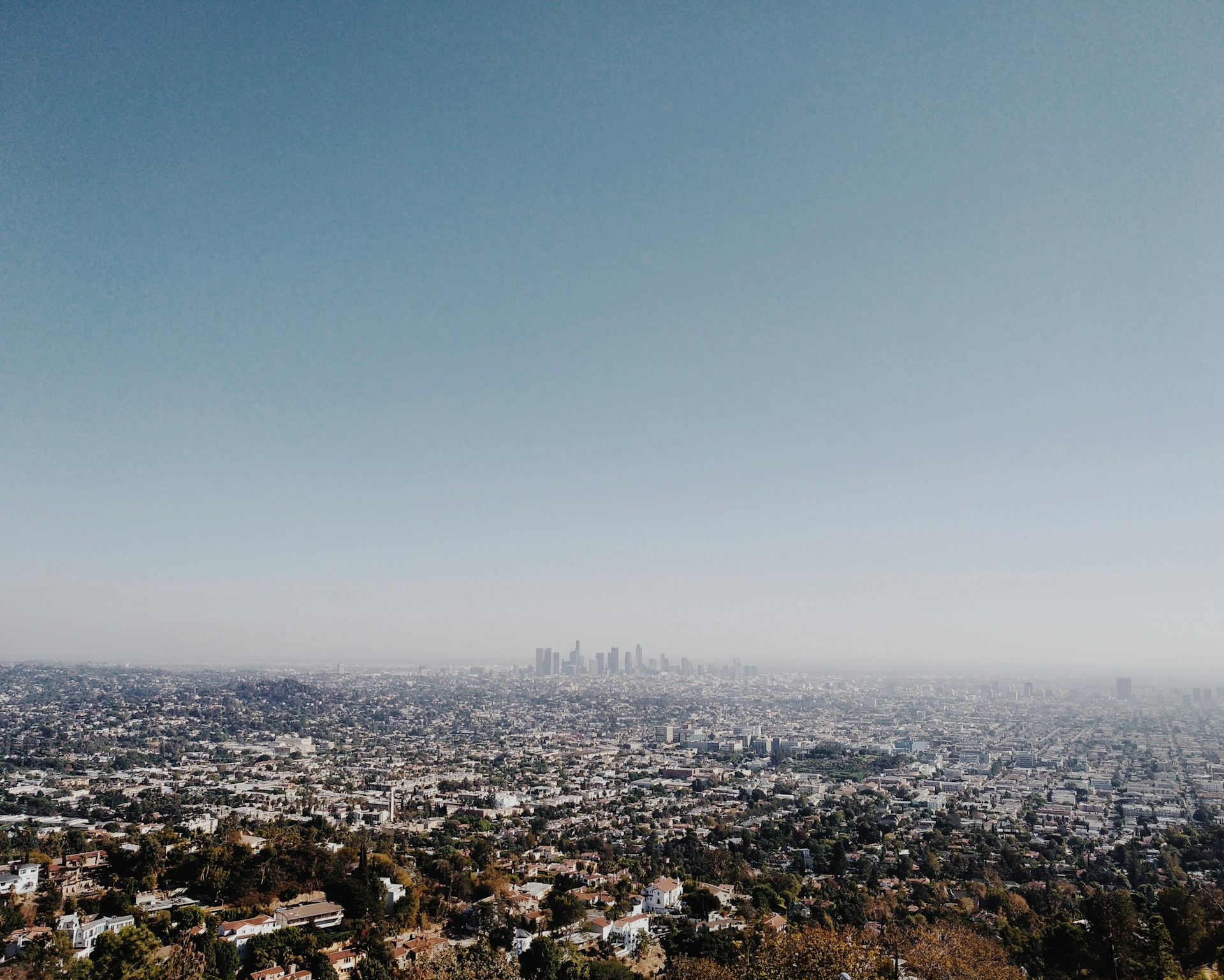 Hazy aerial view of Los Angeles from a hillside, with low residential neighborhoods stretching toward the downtown skyline.